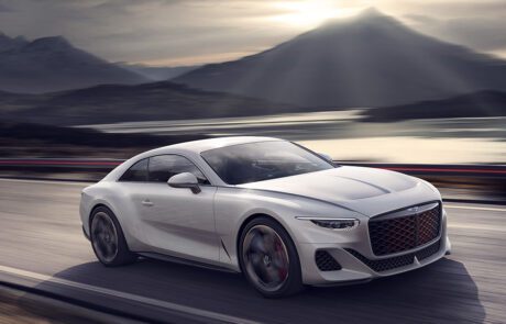 A sleek, silver Bentley Batur Hypercar speeds along a mountain road at dusk. The background features snow-capped mountains, a serene lake with the reflection of the setting sun, and a dramatic sky filled with clouds and sunbeams, all masterfully rendered by a CGI artist. The car's red accents stand out prominently.
