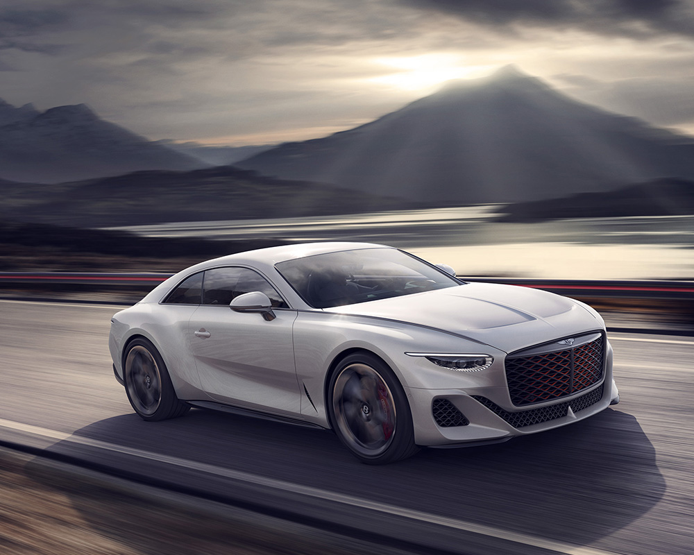 A sleek, silver Bentley Batur Hypercar speeds along a mountain road at dusk. The background features snow-capped mountains, a serene lake with the reflection of the setting sun, and a dramatic sky filled with clouds and sunbeams, all masterfully rendered by a CGI artist. The car's red accents stand out prominently.