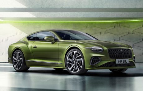 A sleek, green, two-door sports car is parked in a modern, industrial setting. The car features a black roof, silver alloy wheels, and a distinctive grille with a "V44 BML" license plate. The background includes geometric wall designs and neon green lighting.