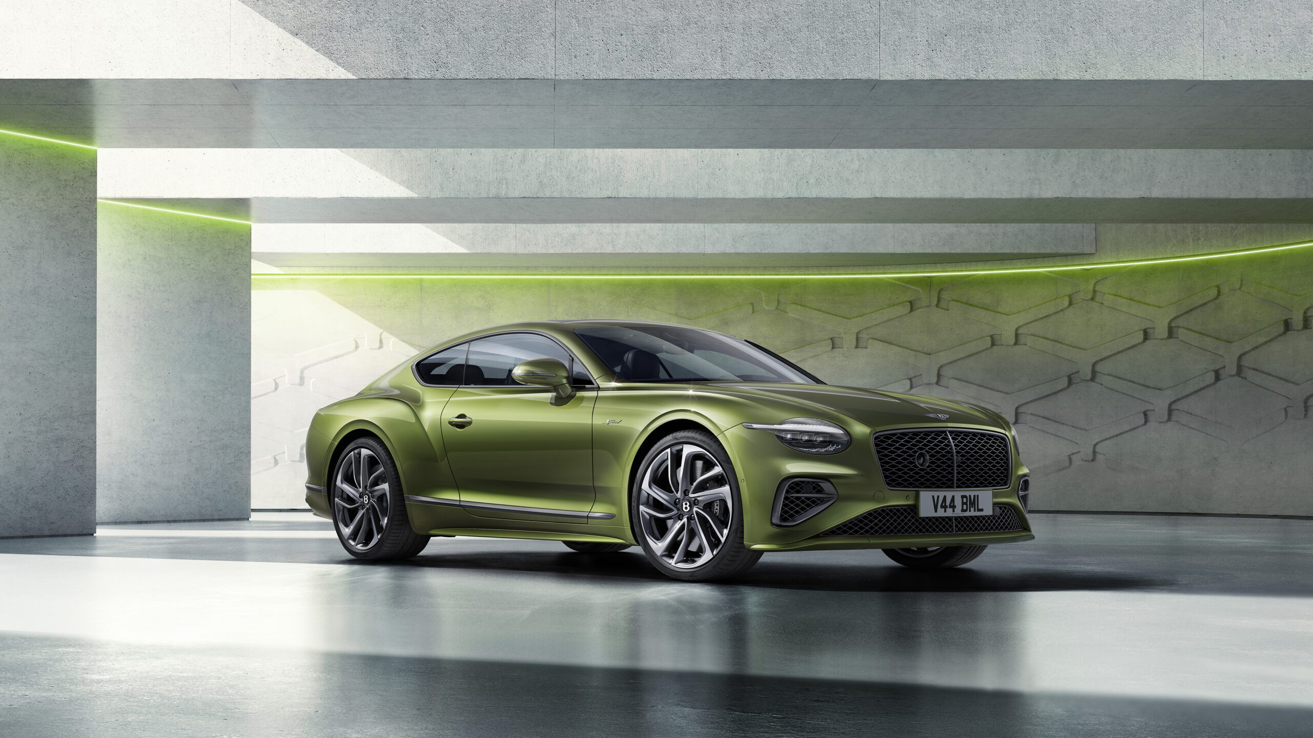 The new Bentley GT Speed is parked indoors in a modern, concrete space with accent lighting. The car features a streamlined design, large alloy wheels, and a sophisticated front grille.