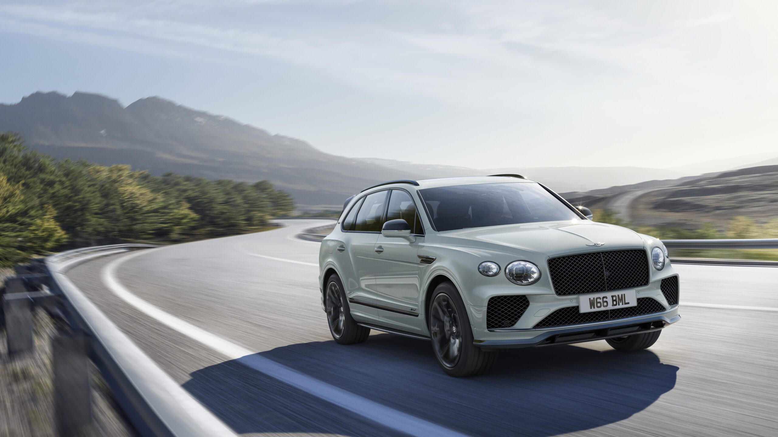 A white Bentley Final Edition Speed12 luxury SUV is driving on a winding mountain road with a scenic backdrop of hills and trees under a partly cloudy sky, captured in stunning CGI.