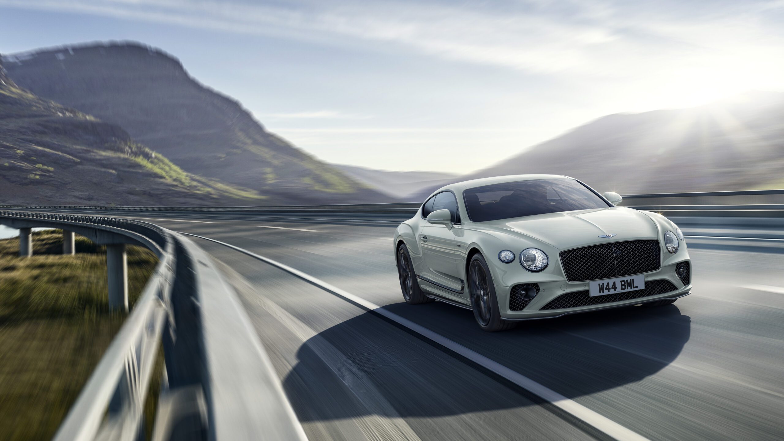 A sleek, silver Bentley Final Edition Speed12 speeds down an expansive, winding highway with guardrails. The CGI car is set against a backdrop of distant mountains and a sunlit sky, conveying a sense of motion and freedom. The license plate reads "M44 BML.