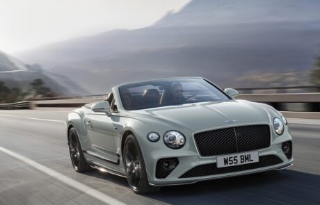 A luxurious light green Bentley Final Edition Speed12 convertible car speeds down a scenic mountain road with a driver at the wheel. The background features blurred mountains and a misty sky, highlighting the car's motion and elegance. The car’s license plate reads "W55 BML.