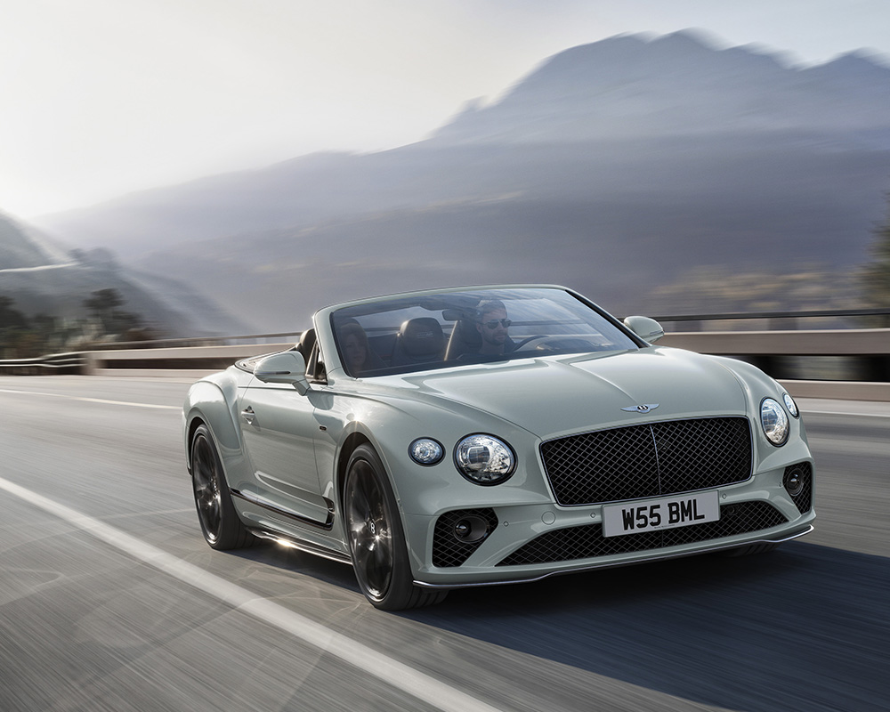 A luxurious light green Bentley Final Edition Speed12 convertible car speeds down a scenic mountain road with a driver at the wheel. The background features blurred mountains and a misty sky, highlighting the car's motion and elegance. The car’s license plate reads "W55 BML.