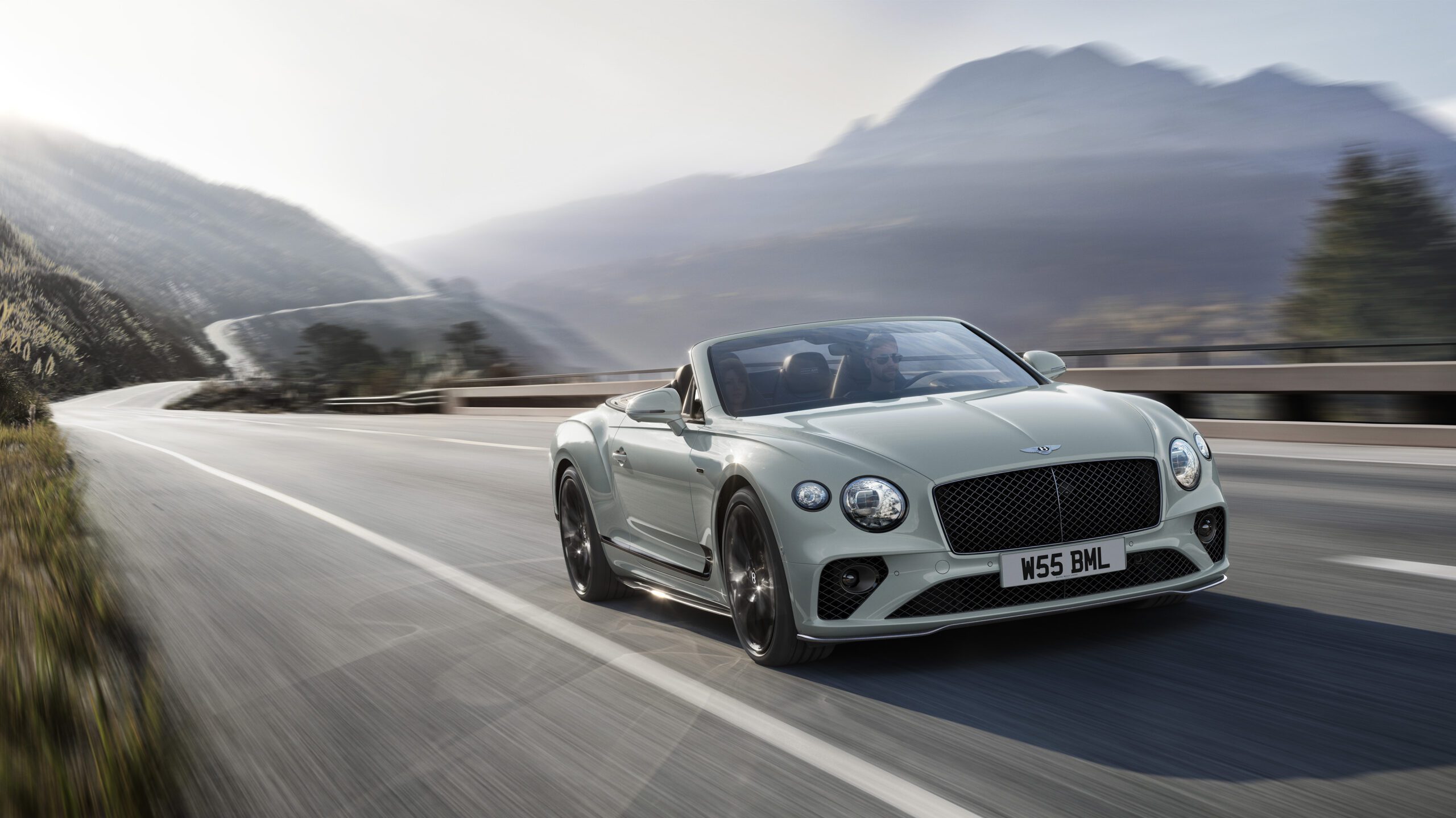 A sleek, light-colored Bentley Final Edition Speed12 convertible sports car speeds along a scenic mountain road. The background features rolling hills, tall trees, and a misty sky, highlighting the car's luxurious design and dynamic performance. CGI perfection makes it look almost ethereal. The license plate reads "W55 BML.