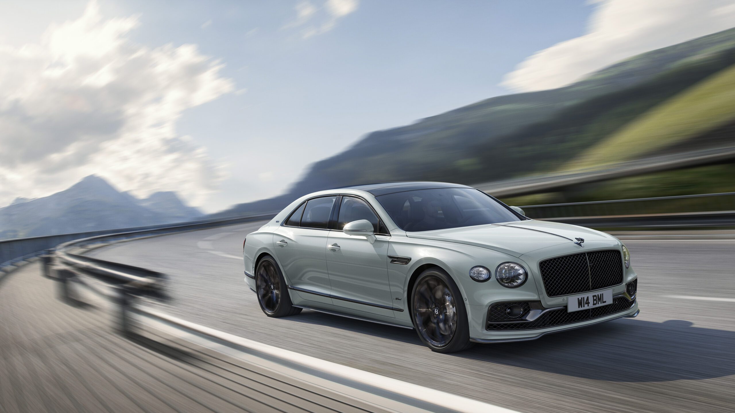 A sleek, light-colored Bentley Final Edition Speed12 luxury sedan with a modern design speeds along a winding mountain road. The vehicle has dark tinted windows and is in motion, creating a dynamic blur effect against the scenic CGI backdrop of mountains under a partly cloudy sky.