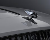 Close-up of a sleek, metallic car hood ornament shaped like a stylized bird with illuminated wings, mounted on the shiny silver-gray hood of a retouched Bentley Flying Spur S. The background is a gradient of dark grays.