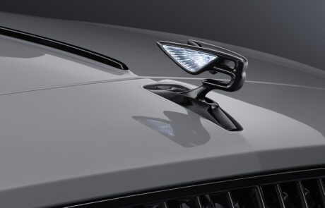 Close-up of a sleek, metallic car hood ornament shaped like a stylized bird with illuminated wings, mounted on the shiny silver-gray hood of a retouched Bentley Flying Spur S. The background is a gradient of dark grays.
