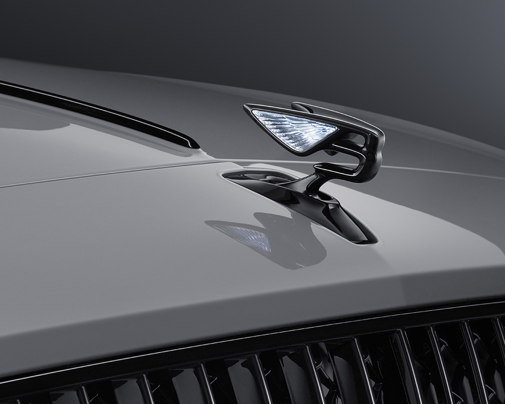 Close-up of a sleek, metallic car hood ornament shaped like a stylized bird with illuminated wings, mounted on the shiny silver-gray hood of a retouched Bentley Flying Spur S. The background is a gradient of dark grays.