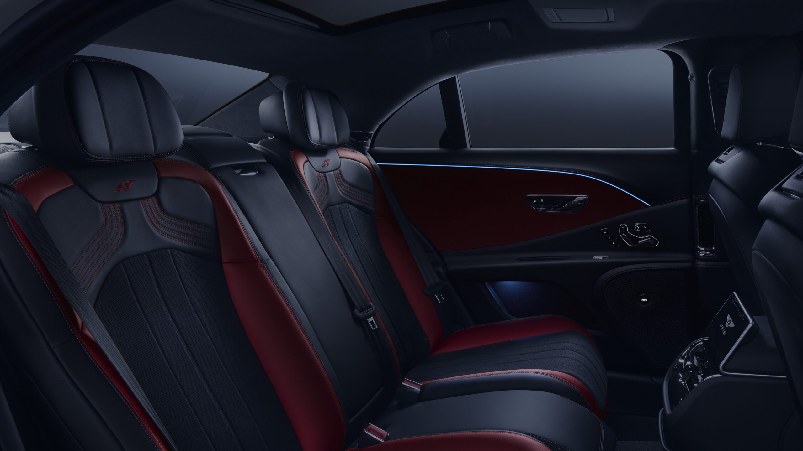 Interior of a Bentley Flying Spur S showcasing the backseat area. The seats are upholstered in black and red leather with intricate stitching. The cabin appears spacious, sophisticated, and well-lit with ambient lighting highlighting the elegant door panels and retouched controls.