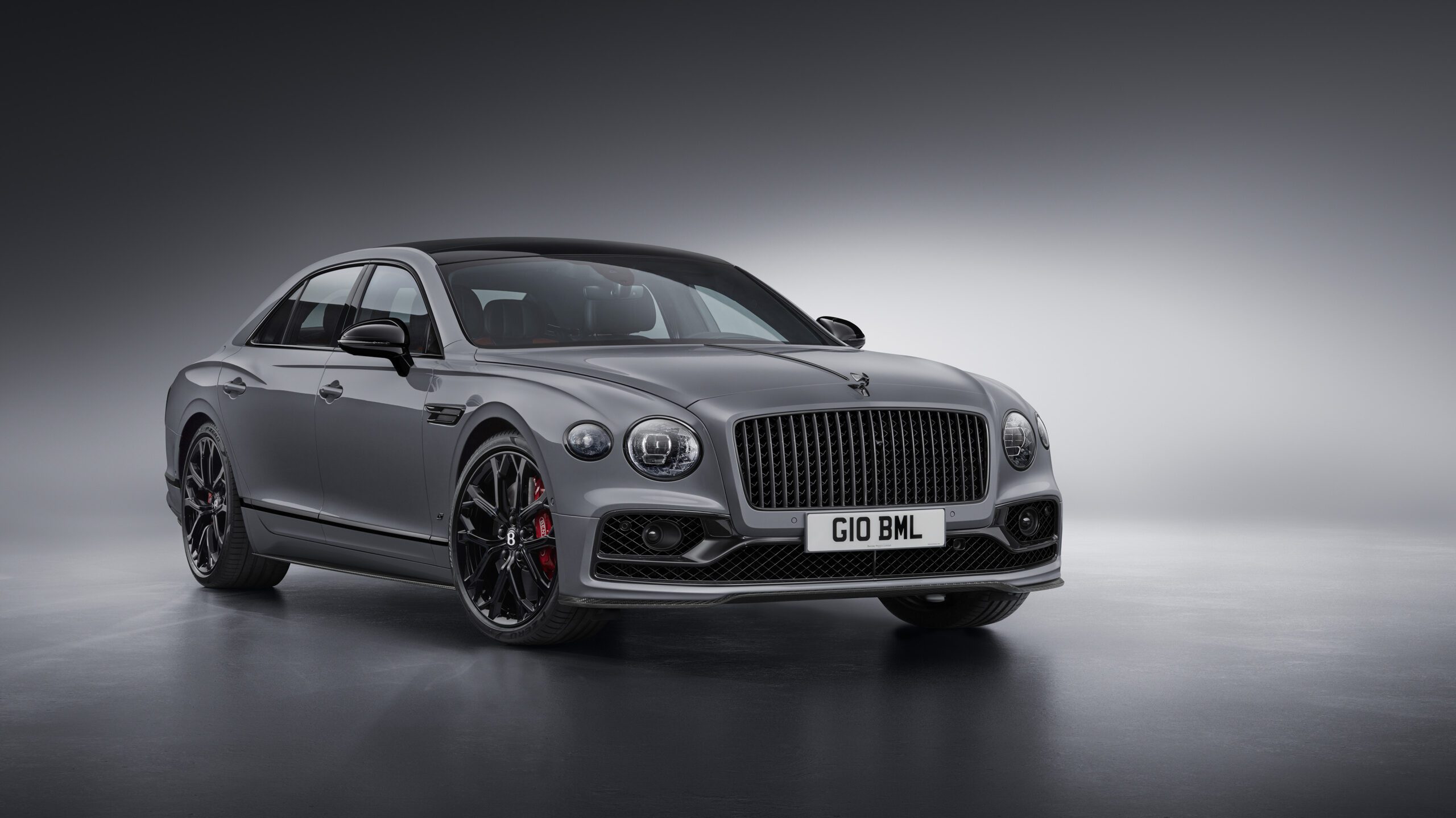 A sleek, silver Bentley Flying Spur S with black accents and red brake calipers is displayed in a modern showroom. The front license plate reads "G10 BML." The car features elegant, aerodynamic curves, large alloy wheels, and a distinctive front grille, all meticulously retouched for perfection.