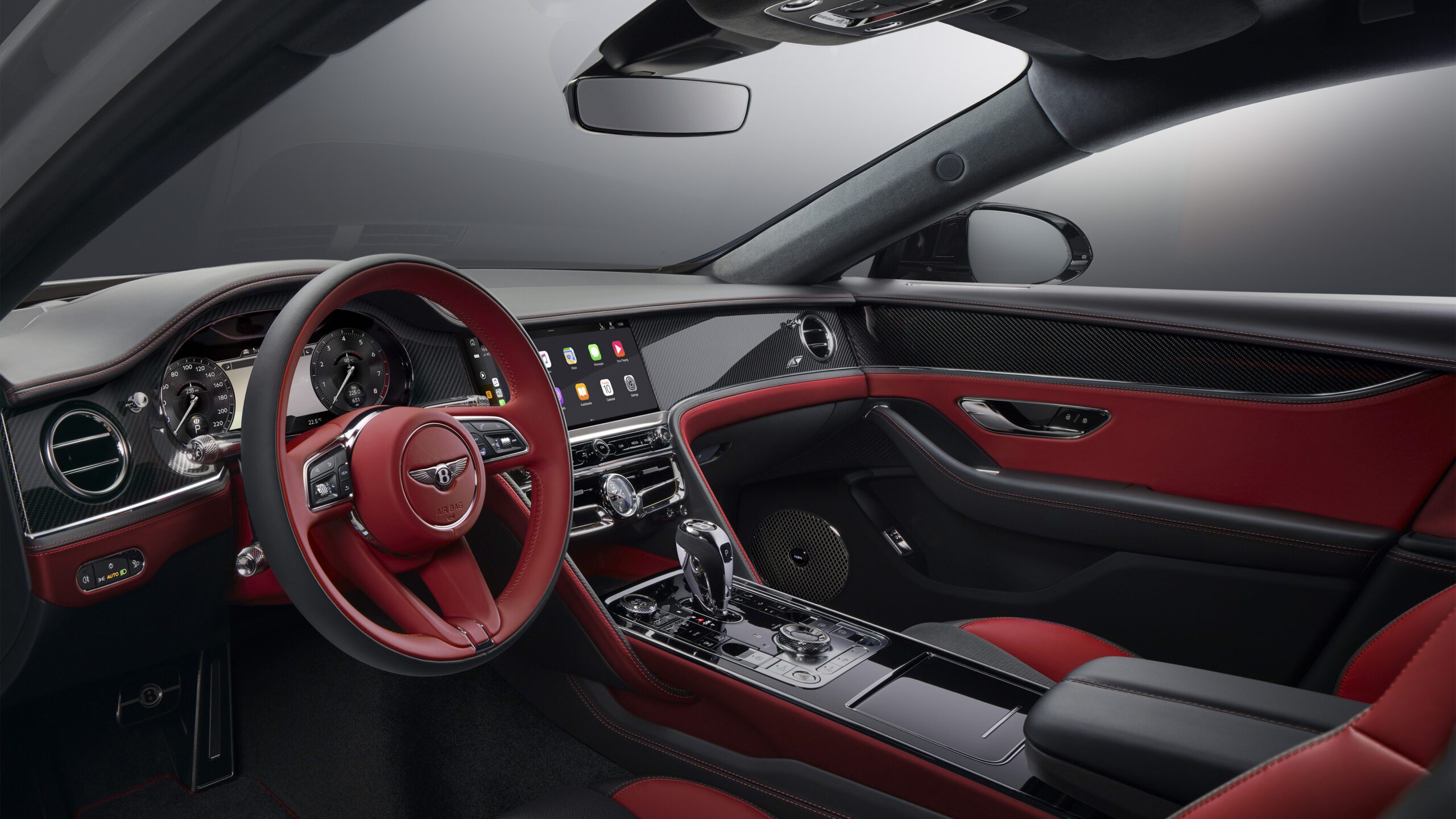 The interior of the Bentley Flying Spur S showcases a retouched luxury ambiance with a red and black color scheme. The dashboard features a digital display, various controls, and a central screen. The steering wheel, seats, and door panels are upholstered in red leather, while the rest of the interior is finished in black with metallic accents.