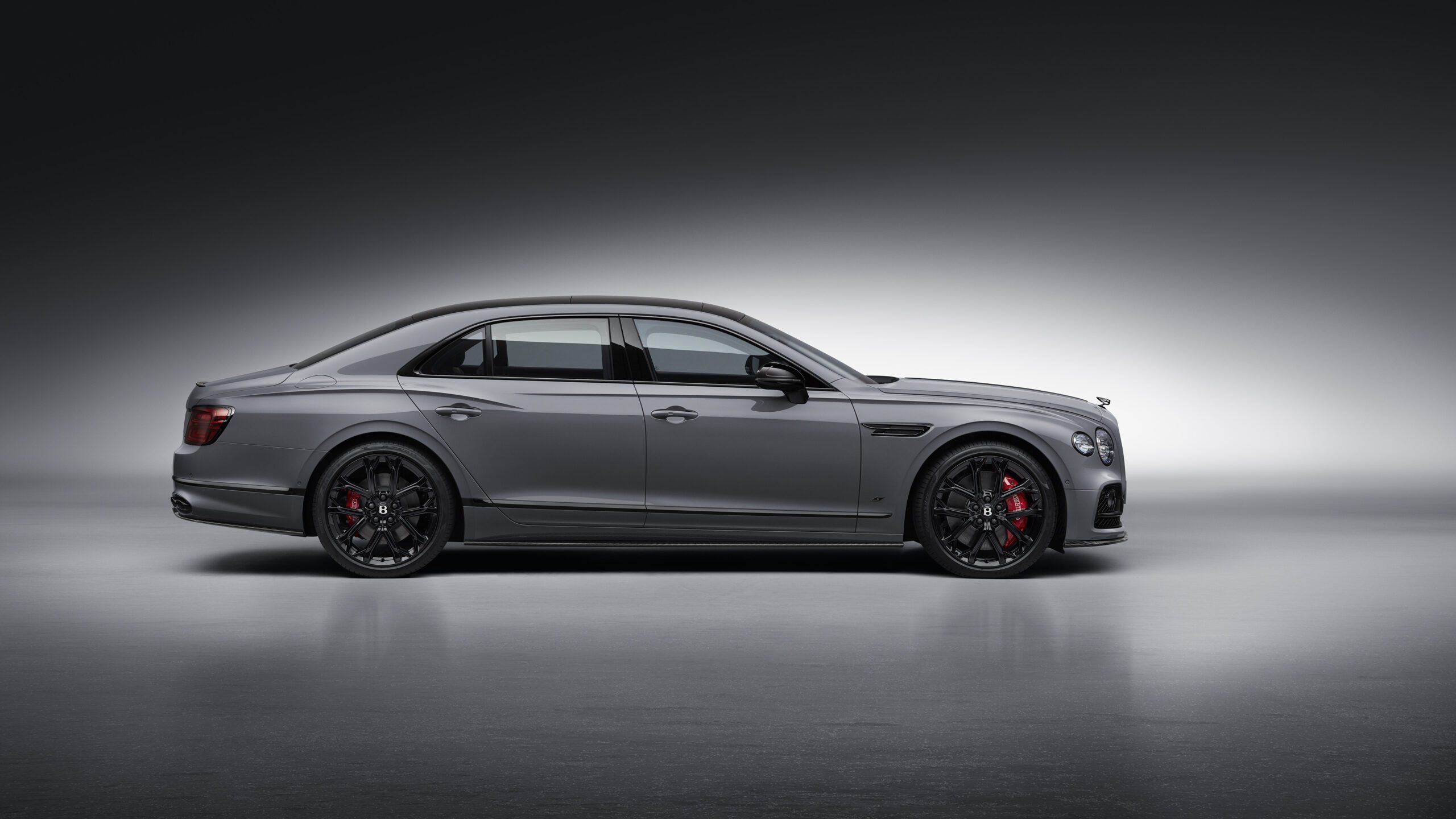 A sleek, metallic gray Bentley Flying Spur S is displayed in a side profile against a gradient gray background. The retouched image highlights the car's black alloy wheels with red brake calipers, tinted windows, and streamlined design.