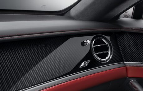 Close-up view of a Bentley Flying Spur S interior featuring a retouched carbon fiber dashboard with red stitching, a circular air vent, and an "S" emblem. The design showcases a sleek, modern aesthetic with attention to detail and craftsmanship.