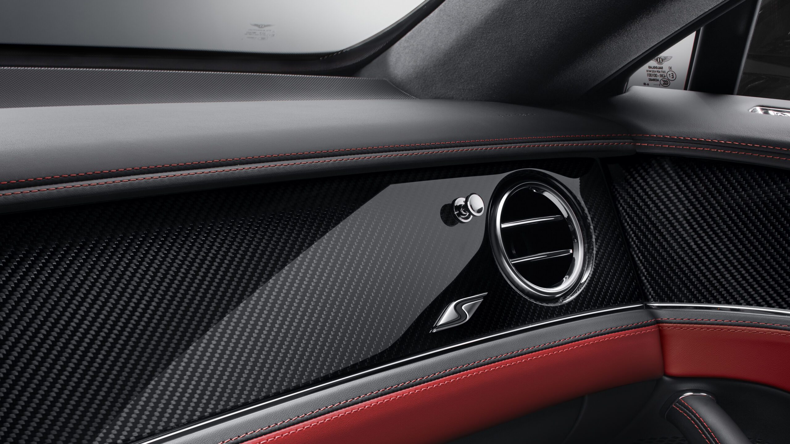 Close-up view of a Bentley Flying Spur S interior featuring a retouched carbon fiber dashboard with red stitching, a circular air vent, and an "S" emblem. The design showcases a sleek, modern aesthetic with attention to detail and craftsmanship.