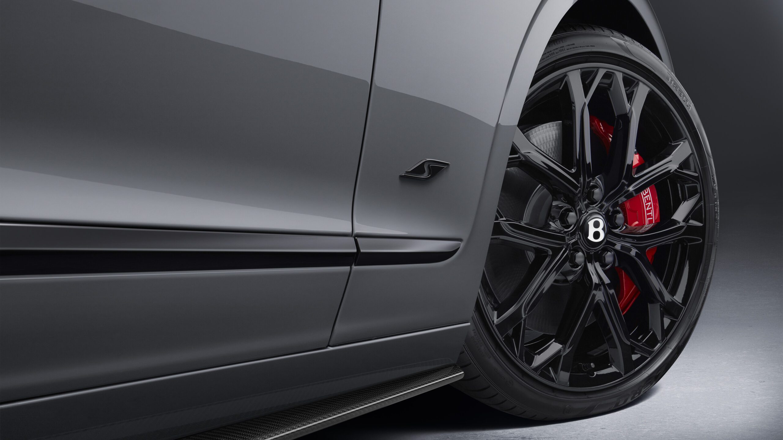 Close-up view of a Bentley Flying Spur S's side, focusing on a sleek, black alloy wheel with a "B" logo in the center and red brake calipers. The glossy gray car body features a subtle badge near the front wheel. The retouched setting is a studio with a dark gradient background.