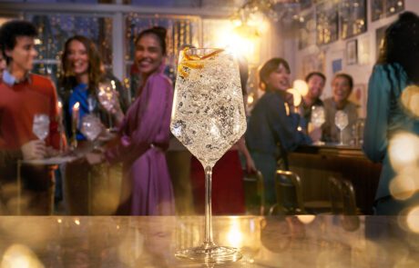 A sparkling Coca Cola royal bliss drink garnished with an orange peel is in focus on a bar counter, while the blurred background shows people enjoying drinks and socializing in a warmly lit bar. Golden bokeh lights add to the festive atmosphere, reminiscent of a scene perfected in a retouchers studio.