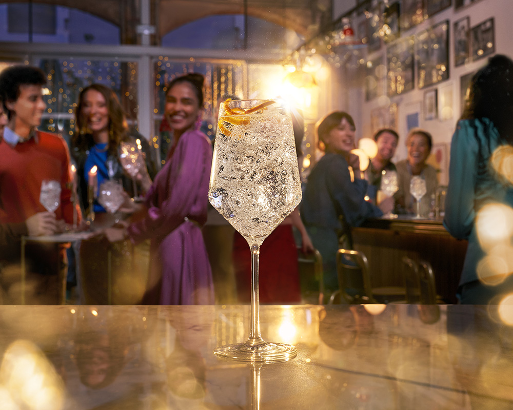 A sparkling Coca Cola royal bliss drink garnished with an orange peel is in focus on a bar counter, while the blurred background shows people enjoying drinks and socializing in a warmly lit bar. Golden bokeh lights add to the festive atmosphere, reminiscent of a scene perfected in a retouchers studio.