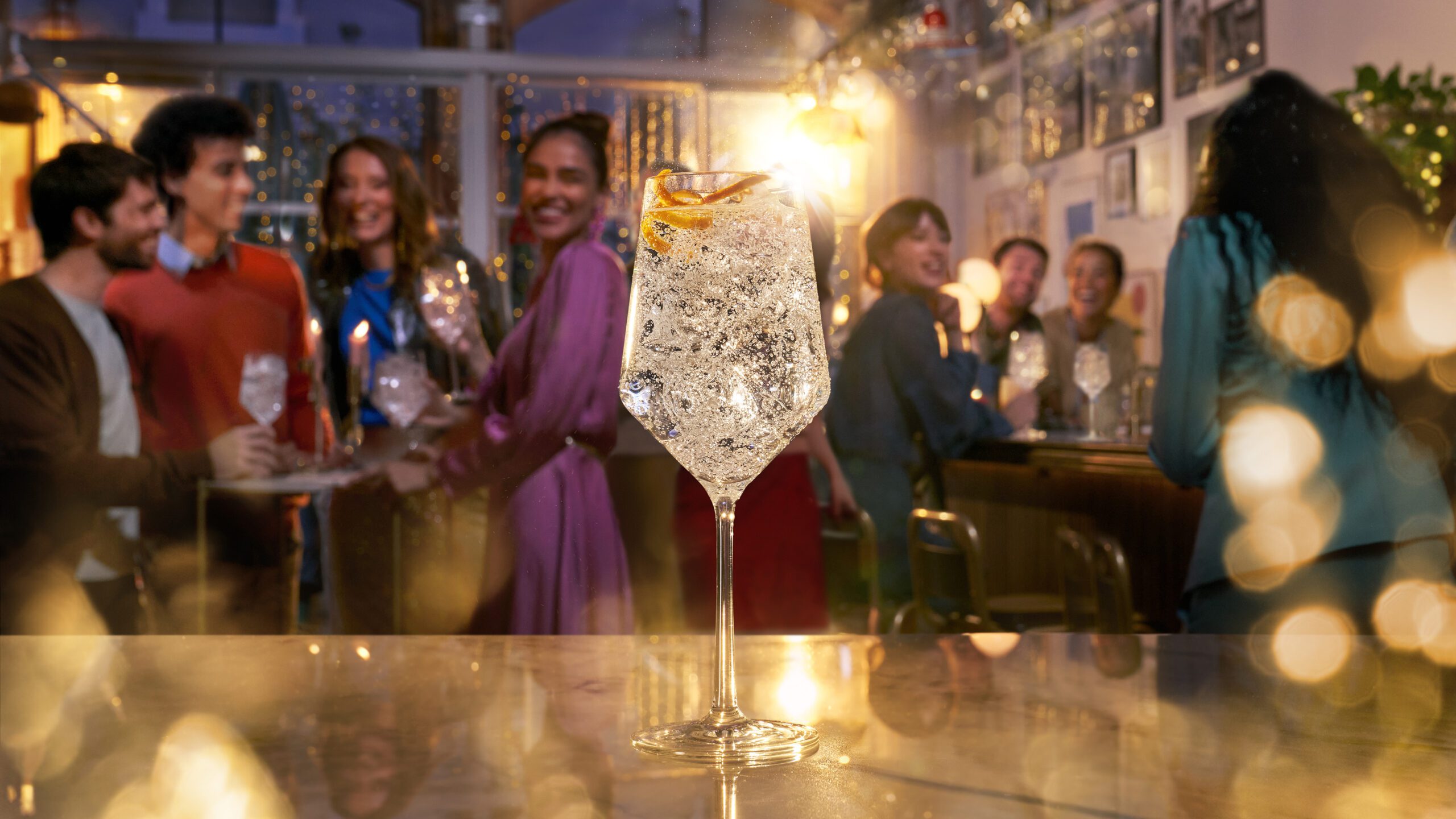 A vibrantly lit bar scene with a large, sparkling cocktail glass in the foreground. In the background, a group of people are joyfully socializing and laughing, creating a festive atmosphere. String lights and decor add to the warm ambiance, reminiscent of a sophisticated retouchers studio.