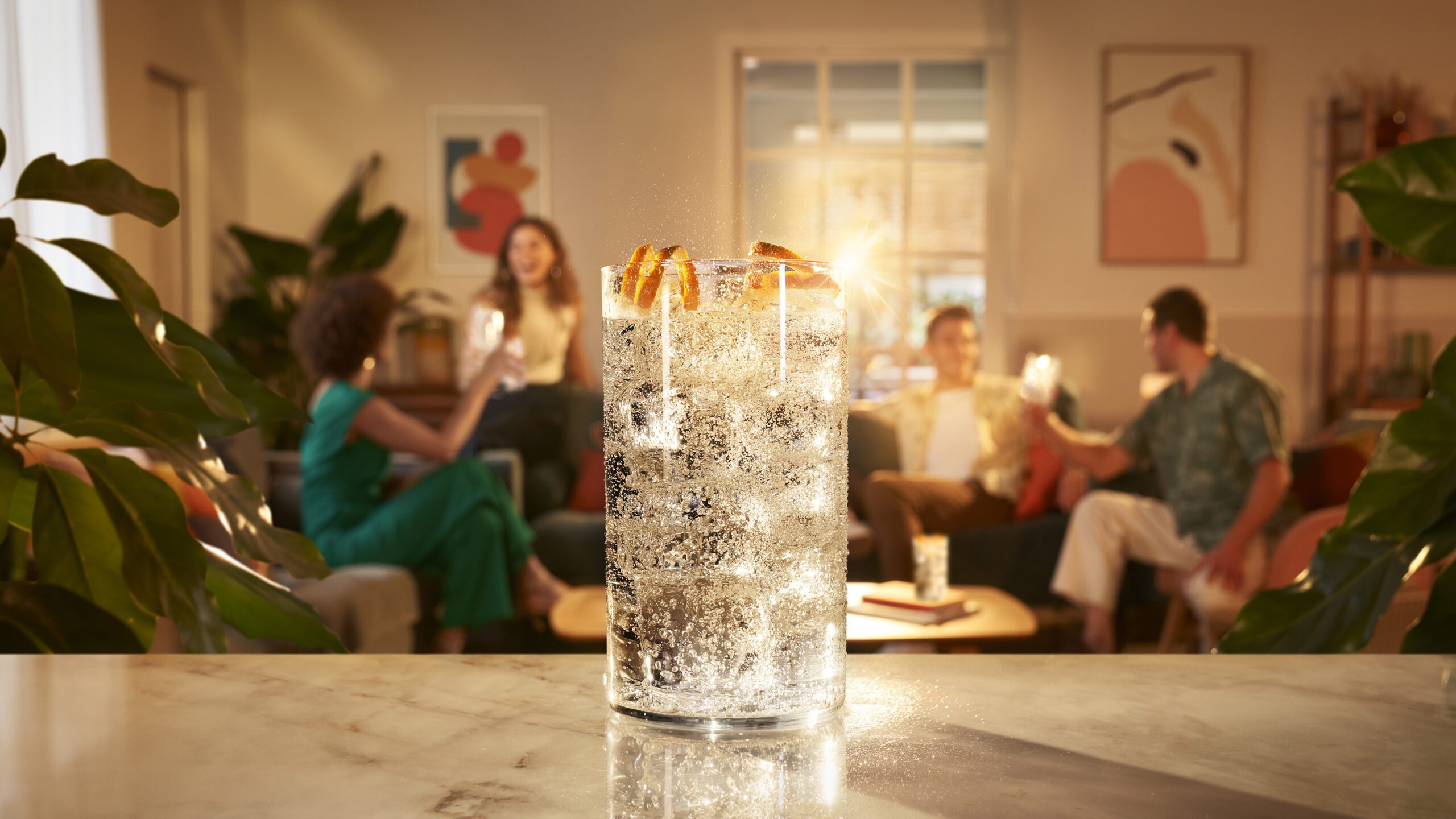 A close-up of a Coca Cola Royal Bliss drink garnished with orange slices on a marble counter. In the background, four people are sitting on couches, engaging in conversation and holding drinks. The setting feels like a cozy, warmly lit living room with modern art and plants, produced by a retoucher's studio.