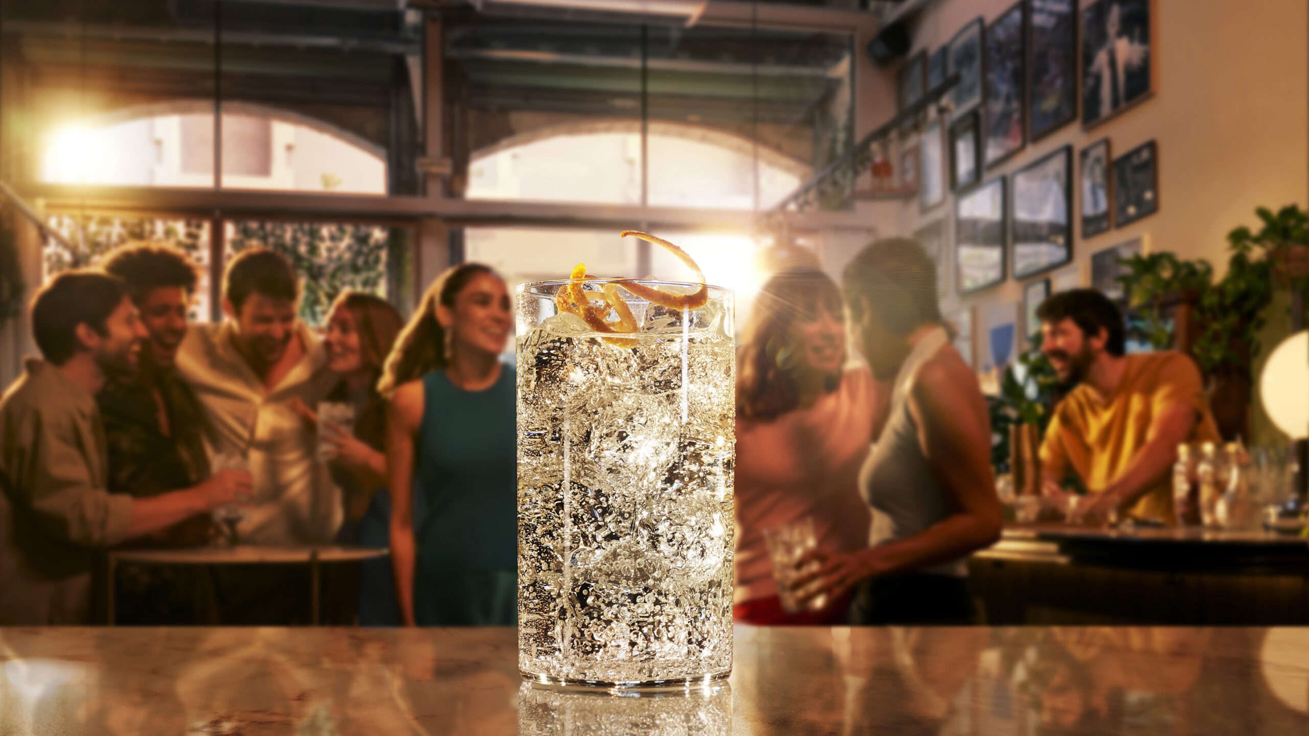 A glass of clear, fizzy cocktail with an orange twist sits on a bar counter. In the blurred background, a group of people are mingling and socializing in a warmly lit bar. The atmosphere appears lively and relaxed, reminiscent of the camaraderie one feels at a retouchers studio.