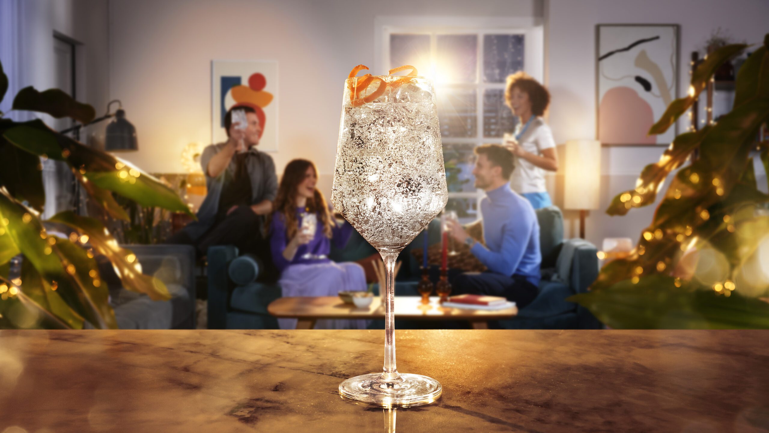 A close-up of a Coca Cola Royal Bliss topped with an orange twist in a glass, placed on a marble counter. In the blurred background, four people are socializing in a cozy, warmly lit living room with modern decor and plants. Perfectly captured as if from a retoucher's studio.