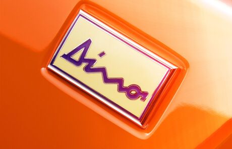 Close-up view of a Ferrari Dino badge with the word "Dino" written in a distinctive script. The badge, detailed by retouchers in London, features a yellow background with a red border and is affixed to an orange surface.