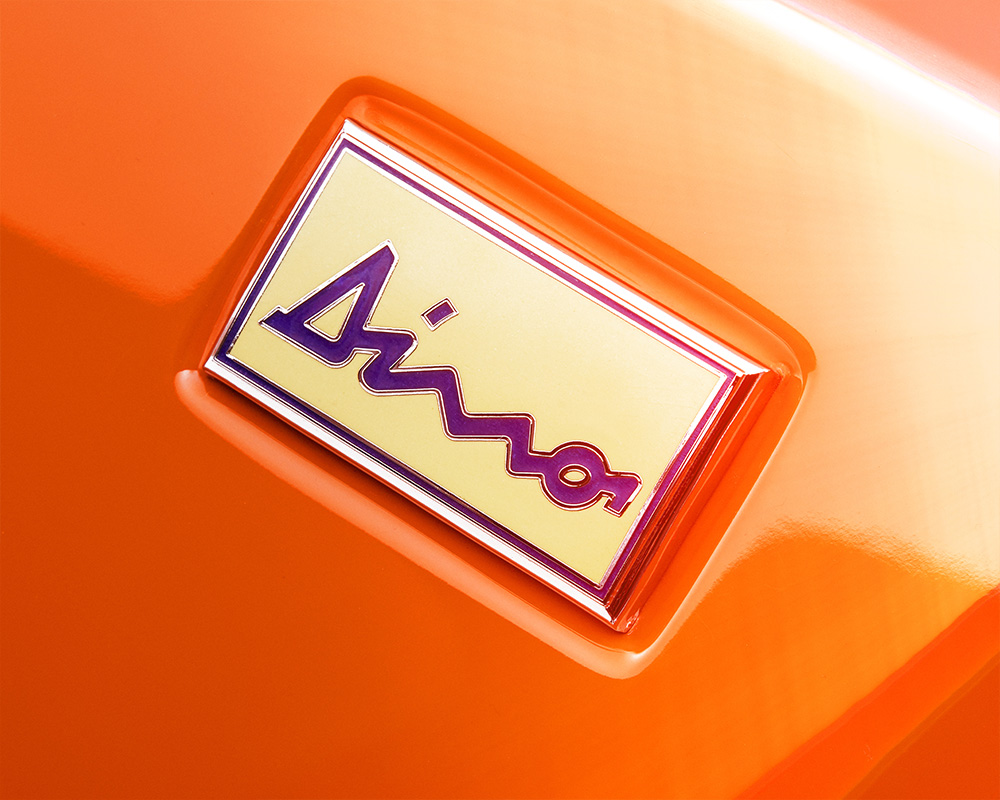 Close-up view of a Ferrari Dino badge with the word "Dino" written in a distinctive script. The badge, detailed by retouchers in London, features a yellow background with a red border and is affixed to an orange surface.