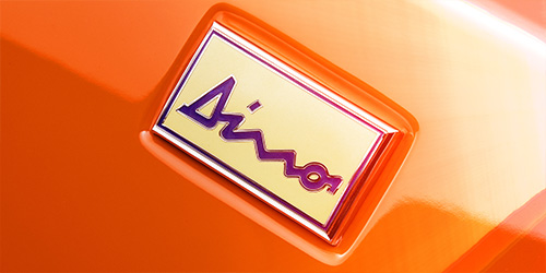 The image shows a close-up of an emblem on an orange surface. The emblem features a yellow background with the word "Dino" in stylish, cursive purple lettering, reminiscent of the iconic Ferrari Dino. This piece could easily be enhanced by expert retouchers in London.