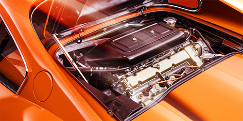 The image shows the engine bay of an orange Ferrari Dino with its hood open. Visible are various engine components, wires, and metallic parts, all neatly arranged within the sleek, glossy exterior of the car—an impeccable capture likely perfected by retouchers in London.