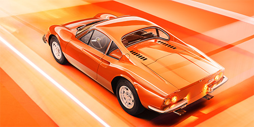 A bright orange Ferrari Dino, with a sleek and aerodynamic design, drives down a road against a matching orange and white gradient background. The car features rounded headlights and distinctive vents on the rear. The image, perfected by retouchers Mustard Post, exudes a sense of speed and style.