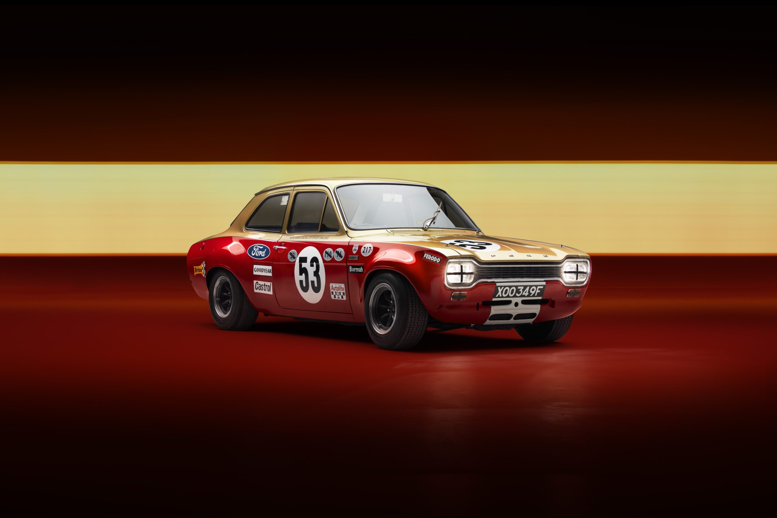 A vintage Ford Escort Mk1 DRVN with a distinctive red and yellow paint job is prominently displayed against a dark, gradient background. The car, featuring the number 53 on the sides and hood, has various sponsorship decals, black wheels, and a classic 1970s design—a true masterpiece for any automotive retoucher.