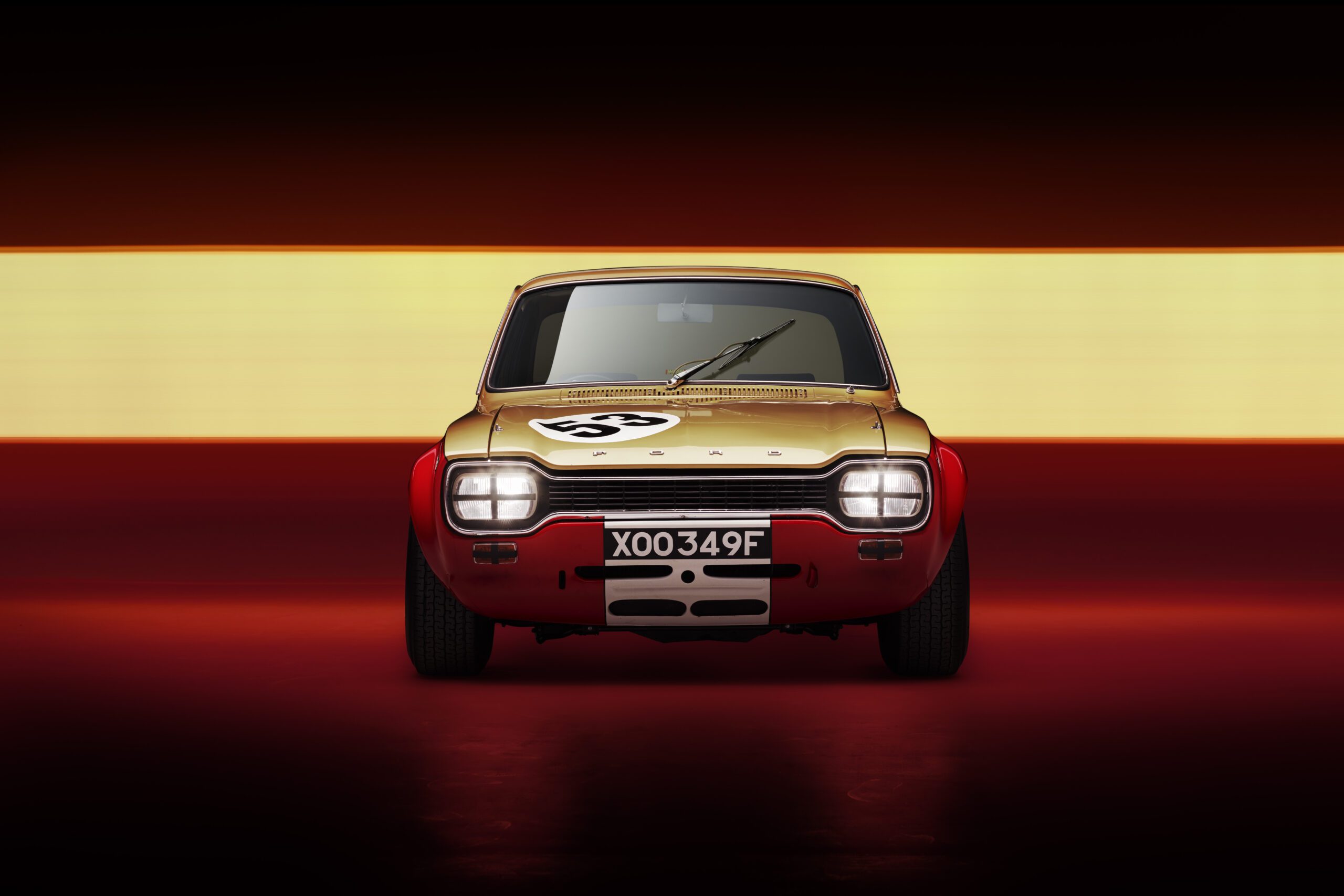 Front view of a classic Ford Escort MK1 DRVN racing car with the number 55 on the hood, showcased against a horizontal yellow light strip on a dark background. The automotive retoucher highlights its distinctive red and gold color scheme and prominent headlights, with license plate XOO349F.