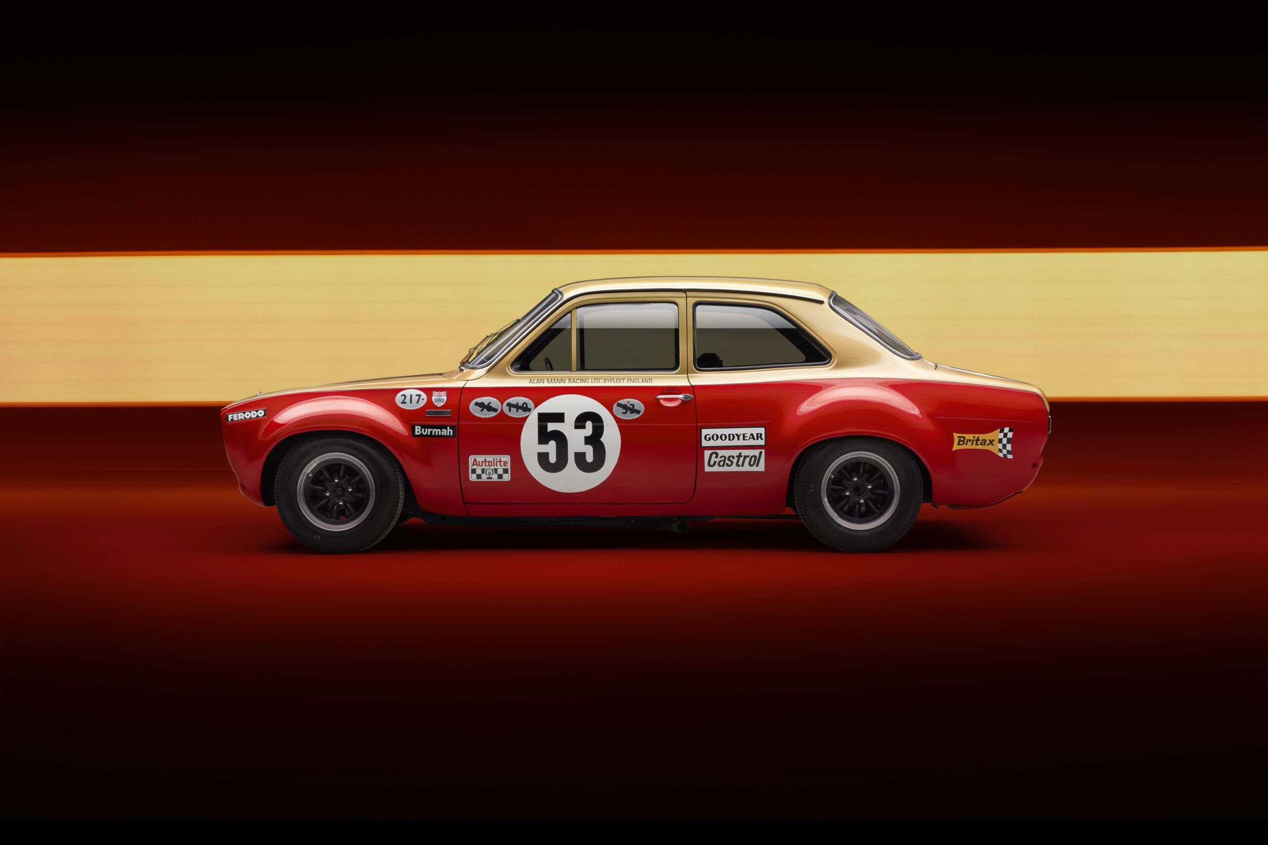 A vintage red Ford Escort Mk1 DRVN race car with the number 53 on the door is displayed against a red background with a horizontal yellow stripe. The car, possibly enhanced by an automotive retoucher, is adorned with various sponsor decals, including Castrol and Dunlop, and features black rims and a classic design.