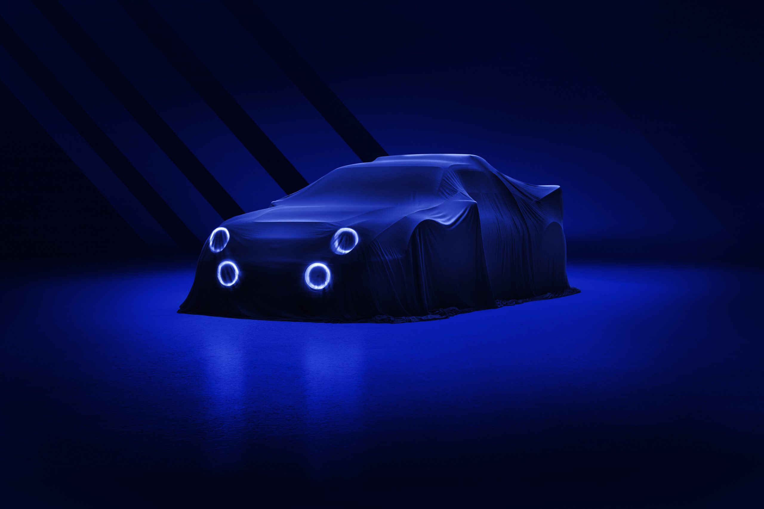 A Ford RS2000 DRVN, covered by a dark cloth, is illuminated by blue lights in an otherwise dark setting. The car retoucher's work highlights the glowing circular headlights and side mirrors, hinting at the vehicle's shape beneath the cover and creating a futuristic, mysterious atmosphere.