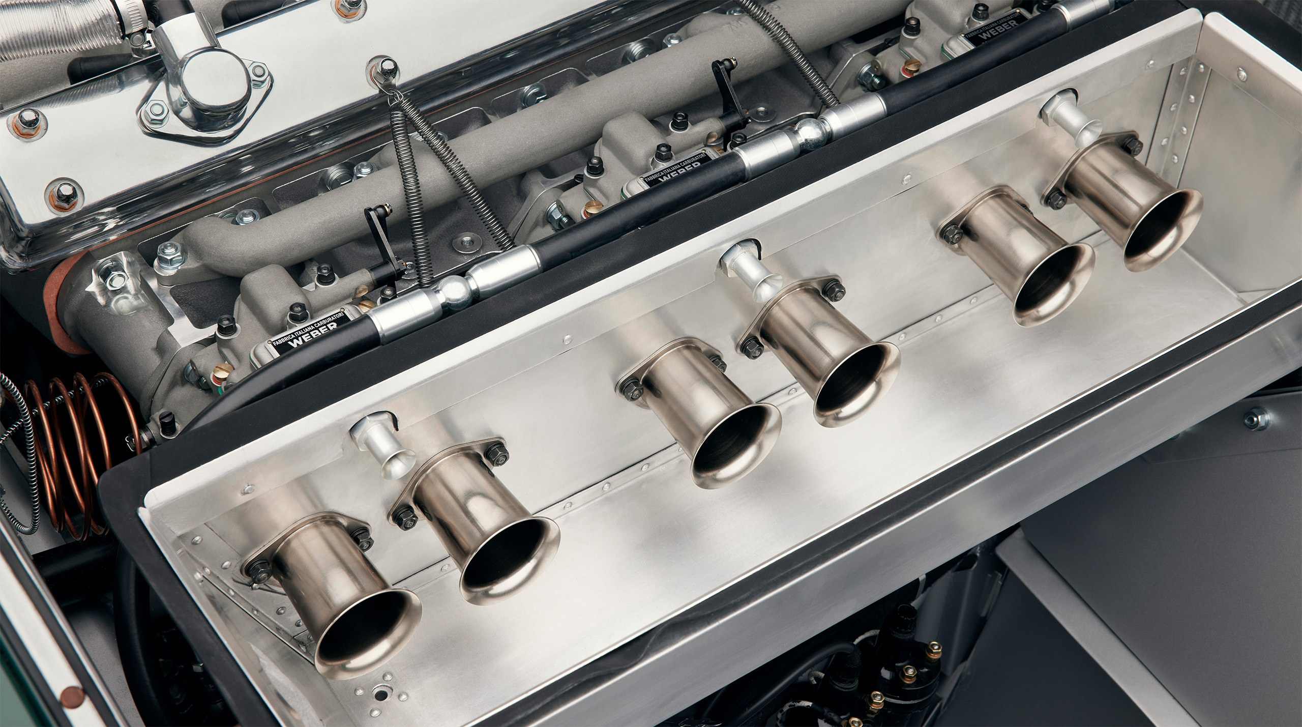 Close-up view of a high-performance car engine, featuring the intake manifold with six polished metal intake tubes. Reminiscent of a Jaguar C-Type, the surrounding components showcase intricate engineering and a metallic finish typical of a racing engine—a perfect subject for any high-end retoucher.