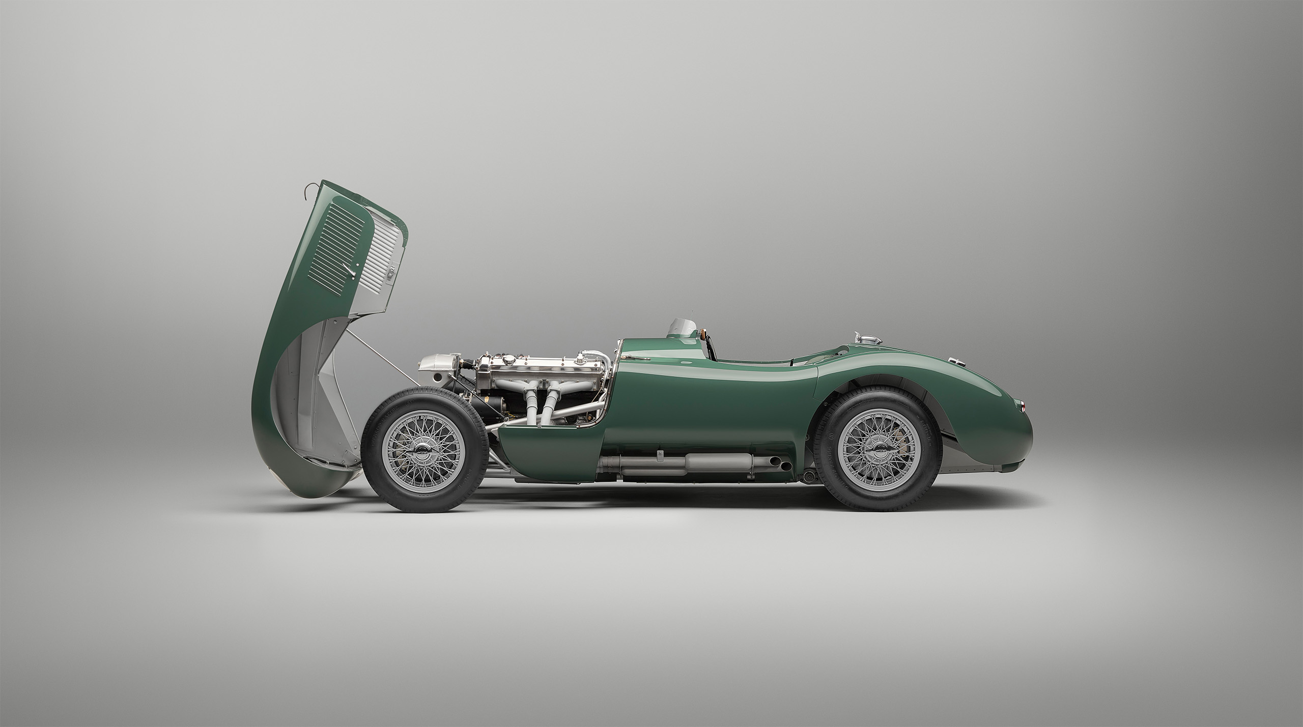 A classic green Jaguar C-Type sports car with its front hood open, revealing the engine. The car is positioned in a side profile view against a neutral background, showcasing its vintage design and detailed mechanical components as expertly captured by a high-end retoucher.