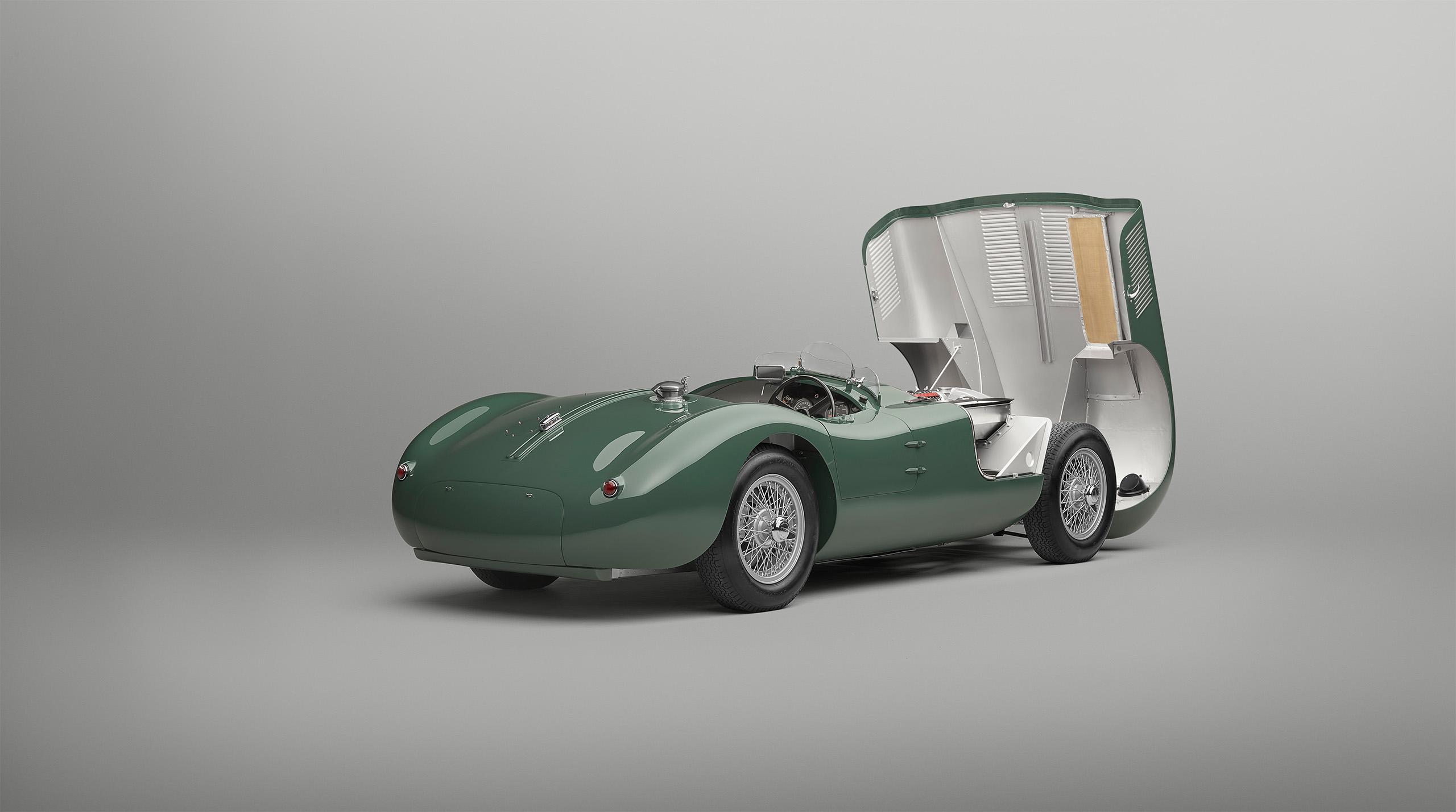 A vintage green Jaguar C-Type with a sleek, aerodynamic design is shown with its rear engine compartment open, revealing intricate mechanical components. The car has wire-spoked wheels and a minimalist cockpit with a small windscreen, all set against a plain background, flawlessly captured by a high-end retoucher.