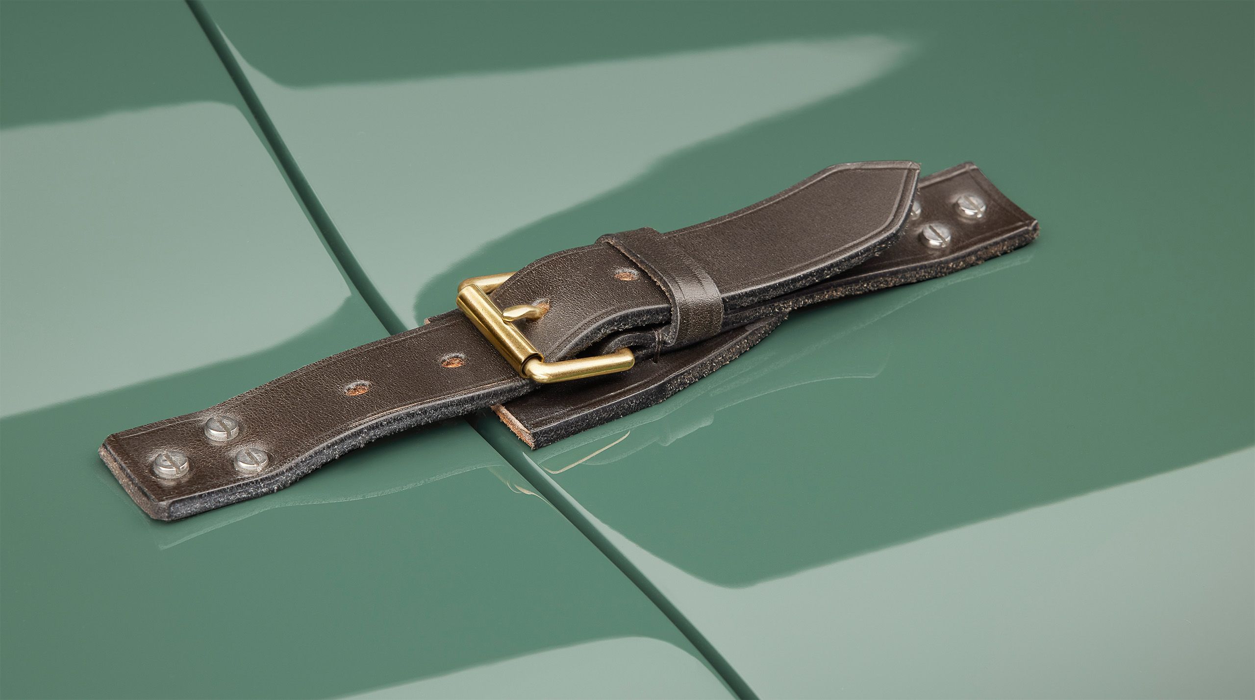 A close-up of a Jaguar C-Type's hood featuring a dark leather strap with a brass buckle. The high-end retoucher has meticulously highlighted the way the strap secures to the green chassis with metal rivets, enhancing its classic design.