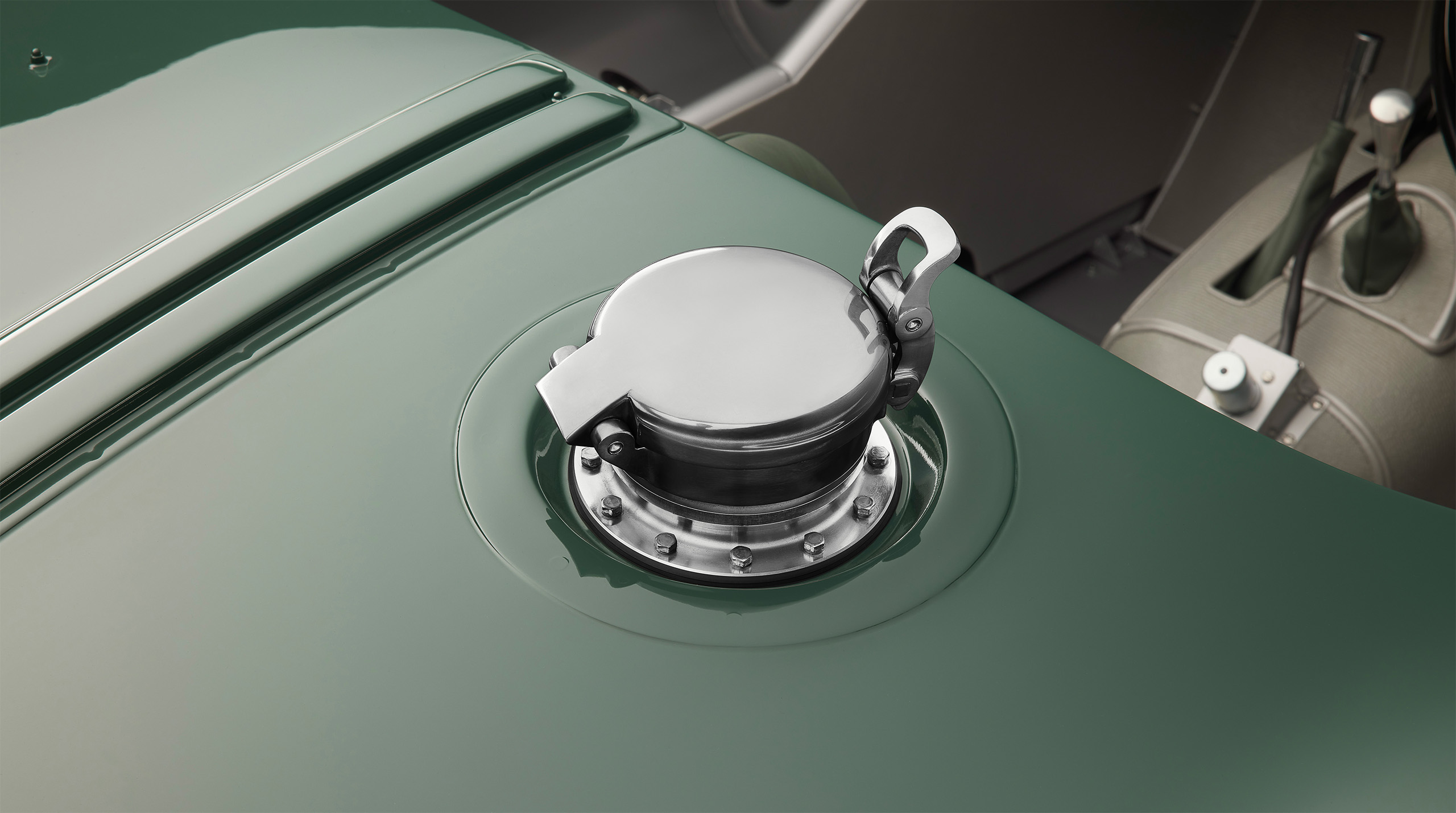 Close-up of the fuel cap on a vintage green car. The polished, metallic cap with a locking mechanism is centrally mounted on a sleek curved surface. A high-end retoucher's touch brings out the details, reminiscent of a Jaguar C-Type. The background reveals part of the car's green and gray interior.