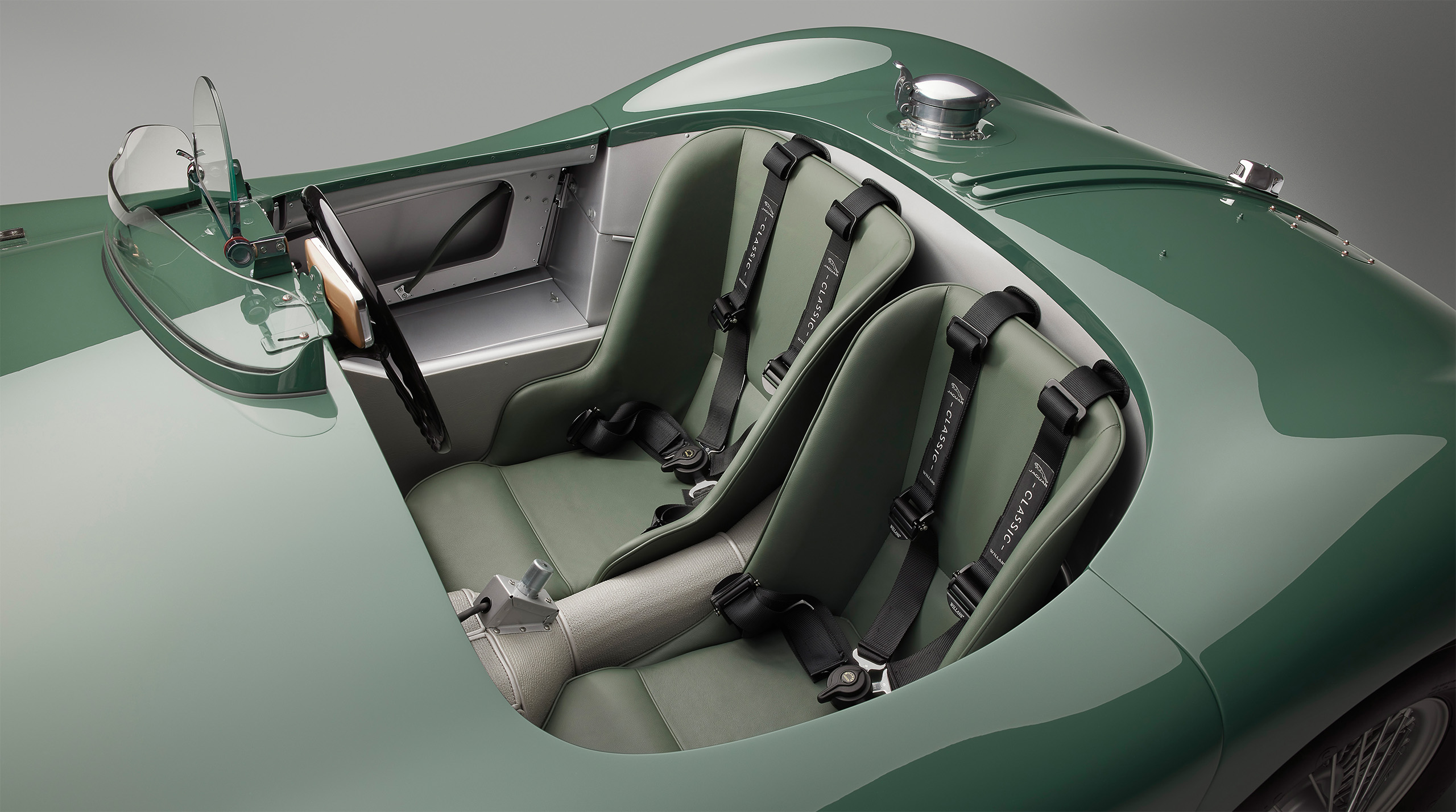 A top view of a vintage green Jaguar C-Type racing car's interior. It features two bucket seats with four-point harnesses upholstered in matching green. The cockpit includes a sleek, minimalist dashboard, emphasizing its racing heritage—perfect for a high-end retoucher's attention to detail.