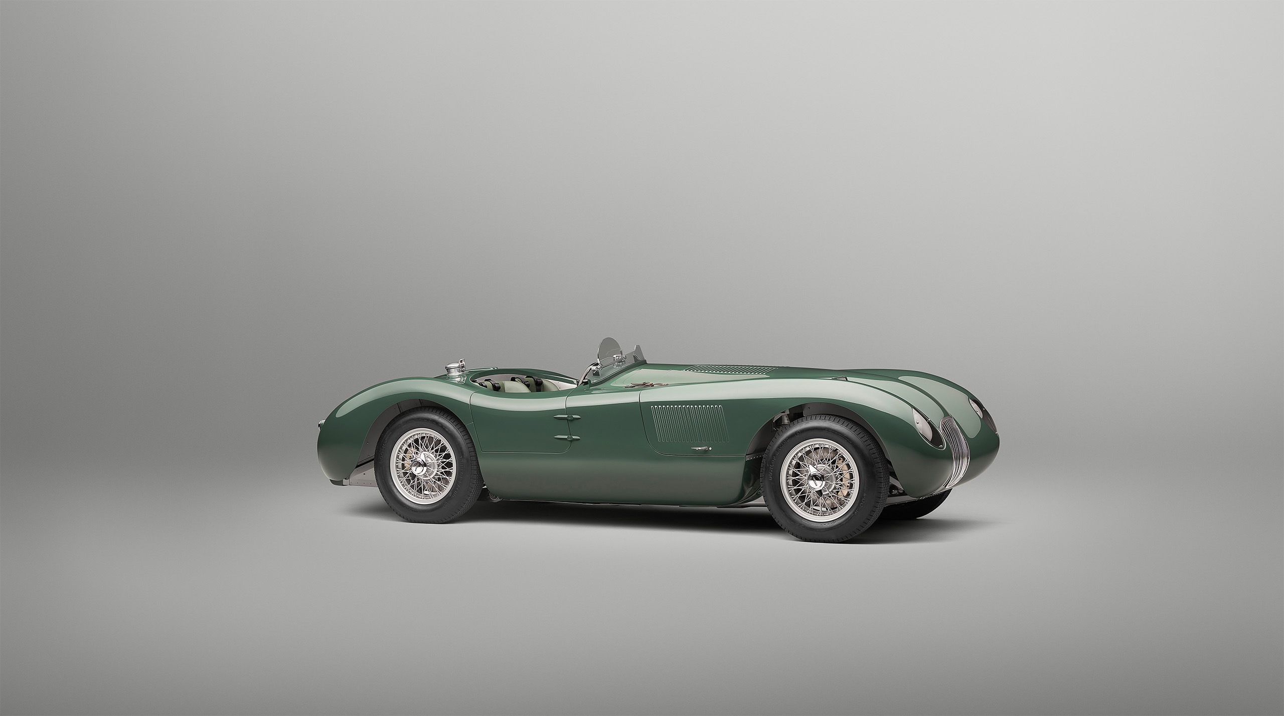 A classic green Jaguar C-Type sports car with a sleek, low-slung design, wire-spoke wheels, and an open cockpit. The car is viewed from the side against a plain, light gray background, highlighting its aerodynamic shape and vintage styling, expertly captured by a high-end retoucher.