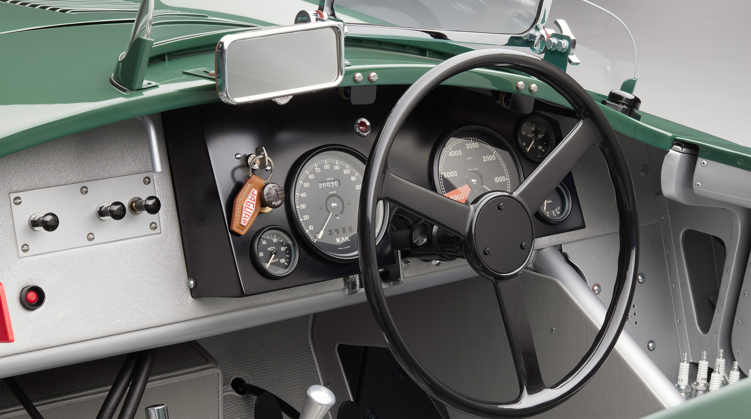 Close-up view of the dashboard and steering wheel of a vintage car. The dashboard features several analog gauges, switches, and buttons. The steering wheel is black with a simple design, reminiscent of a Jaguar C-Type. A keychain with a "MINX" tag hangs from the ignition.