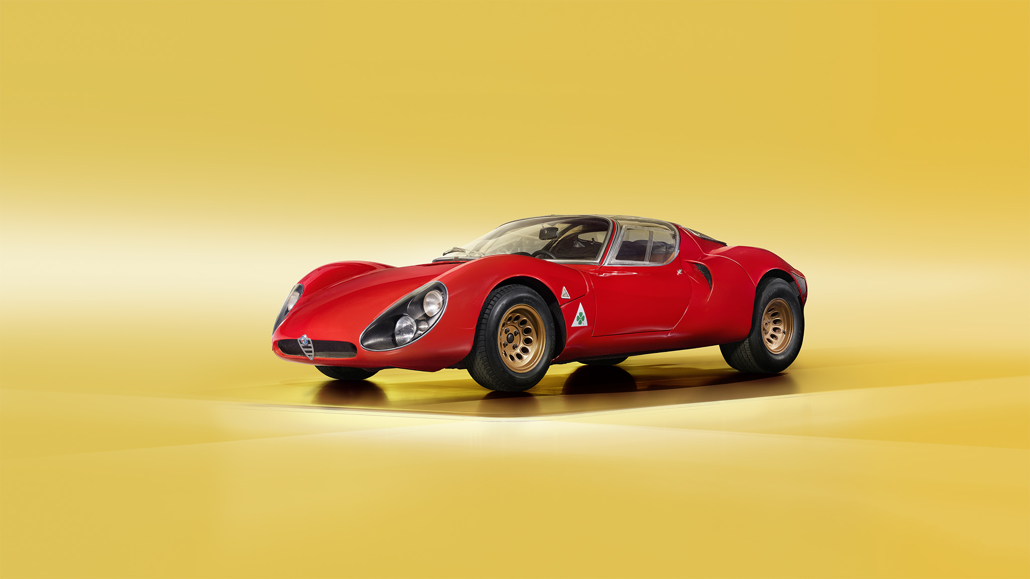 A classic red sports car with sleek curves and a low profile design is displayed in front of a plain yellow background. The Alfa Romeo 33 features round headlights, aerodynamic bodywork, and gold-colored wheels, showcasing a vintage racing aesthetic perfect for a Magneto Magazine cover.