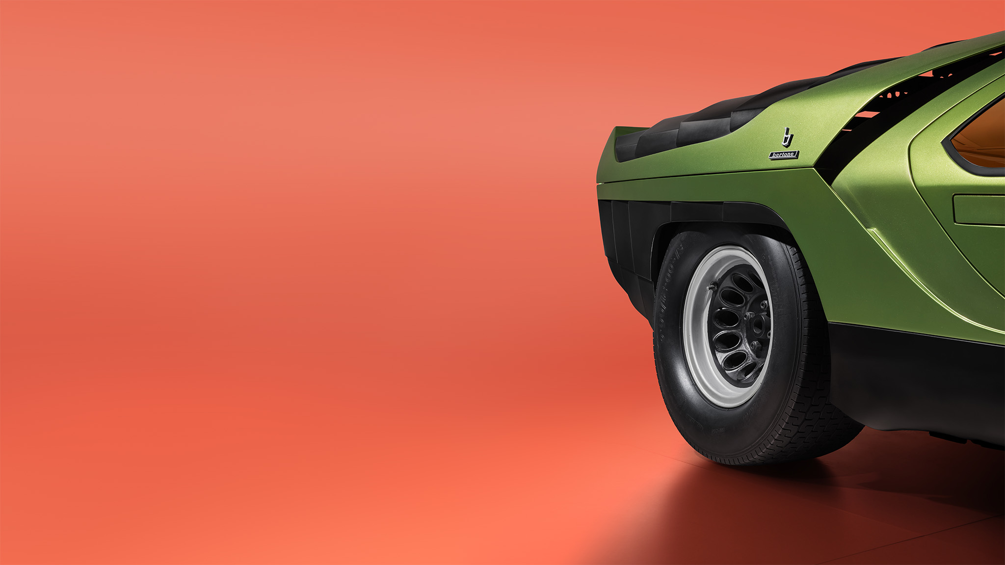 Close-up side view of the rear section of a green classic Alfa Romeo 33 sports car with a distinct angular design, parked on a smooth orange surface. The visible features include the rear wheel, part of the wheel arch with a vent, and part of a tinted window. The background is plain.