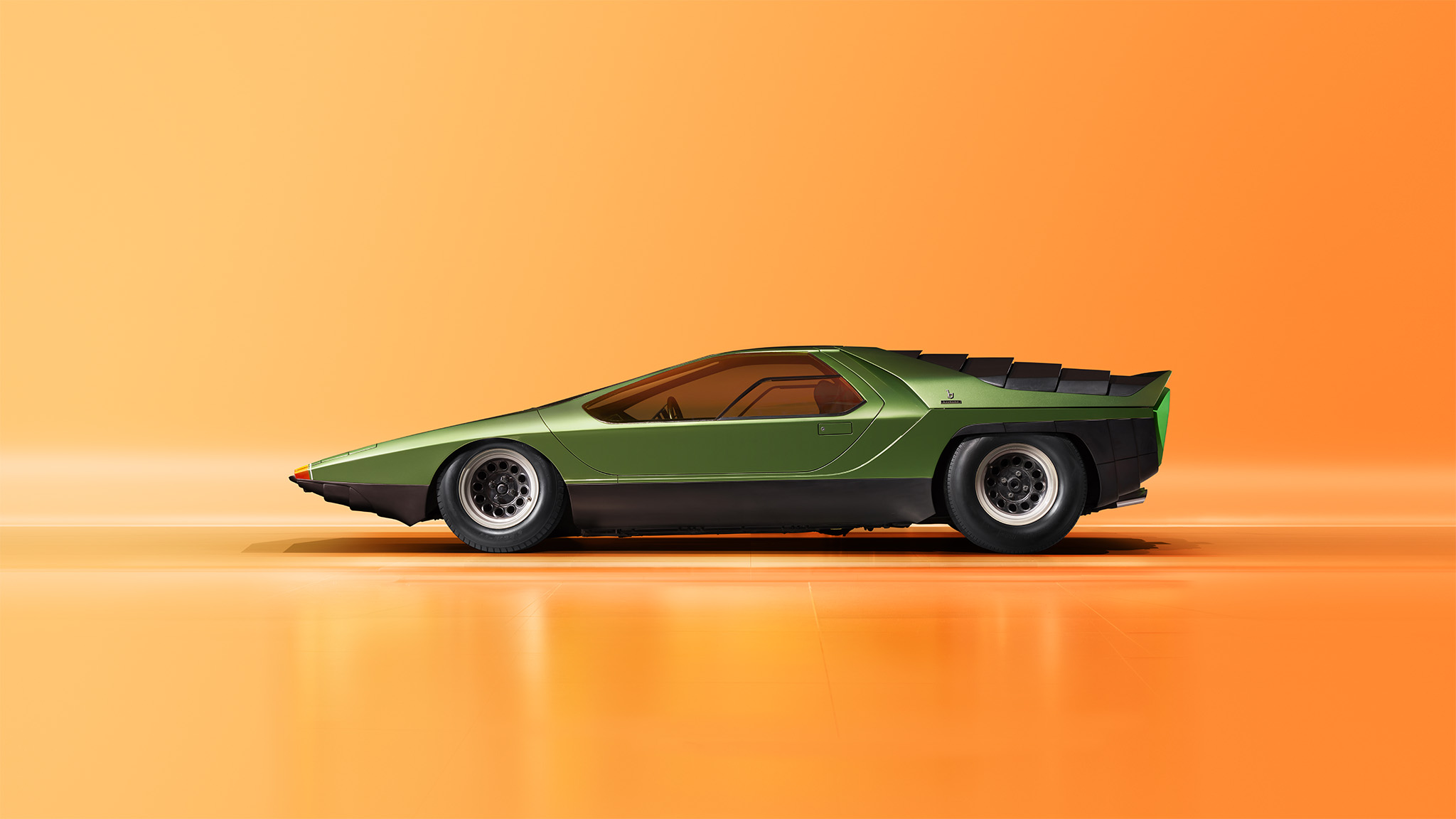 A sleek, retro-futuristic sports car with a low profile and angular design, featuring a green and black color scheme, photographed against a bright orange background. The car has a wedge-shaped nose and large rear wheels, giving it a dynamic, fast look reminiscent of an Alfa Romeo 33.