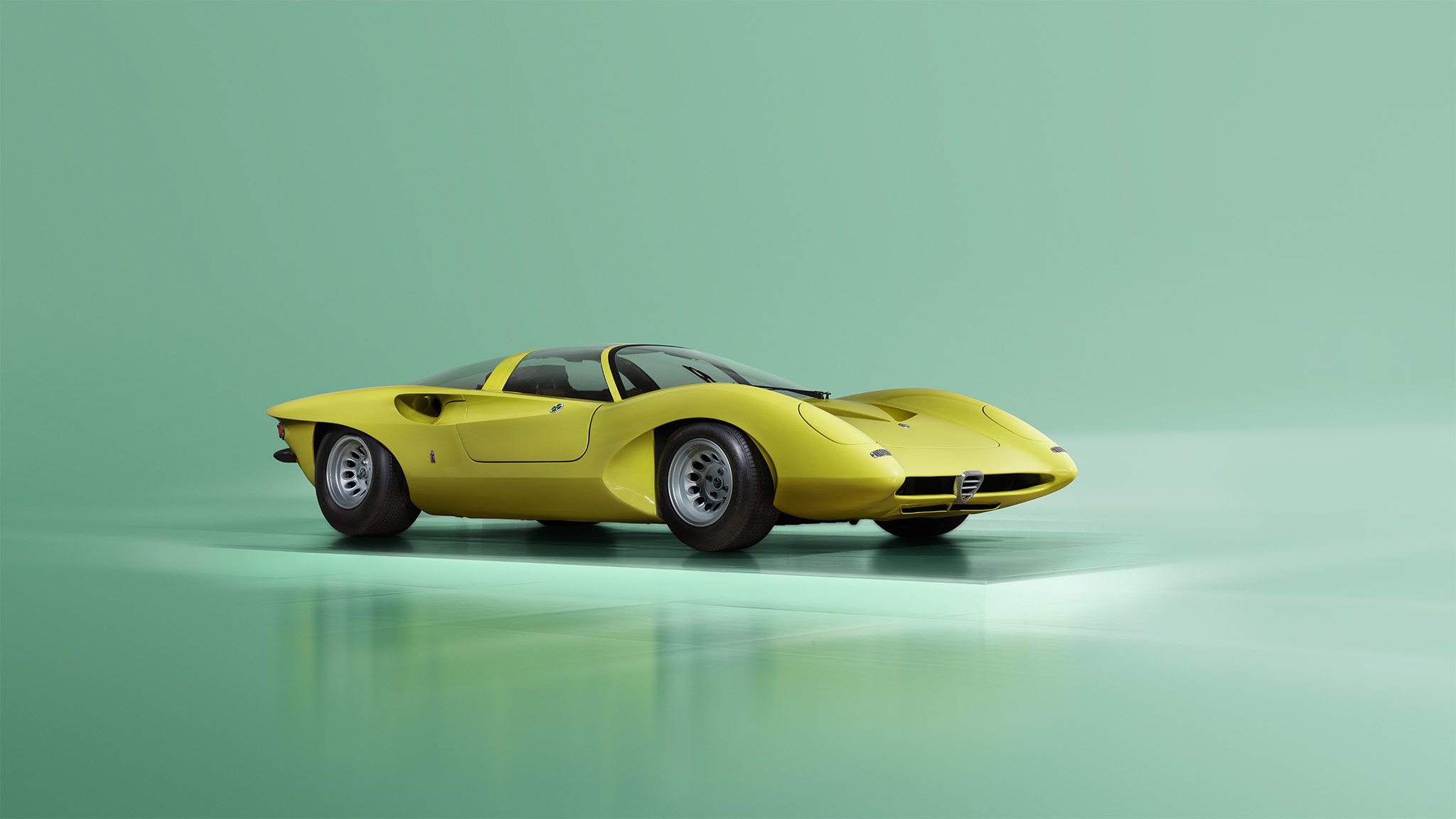 A sleek, yellow Alfa Romeo 33 sports car with a low profile and aerodynamic design is parked on a shiny surface. The background is a gradient of light to dark green. The car features a rounded front, large tires, and a black roof, perfectly captured as if for Magneto Magazine.