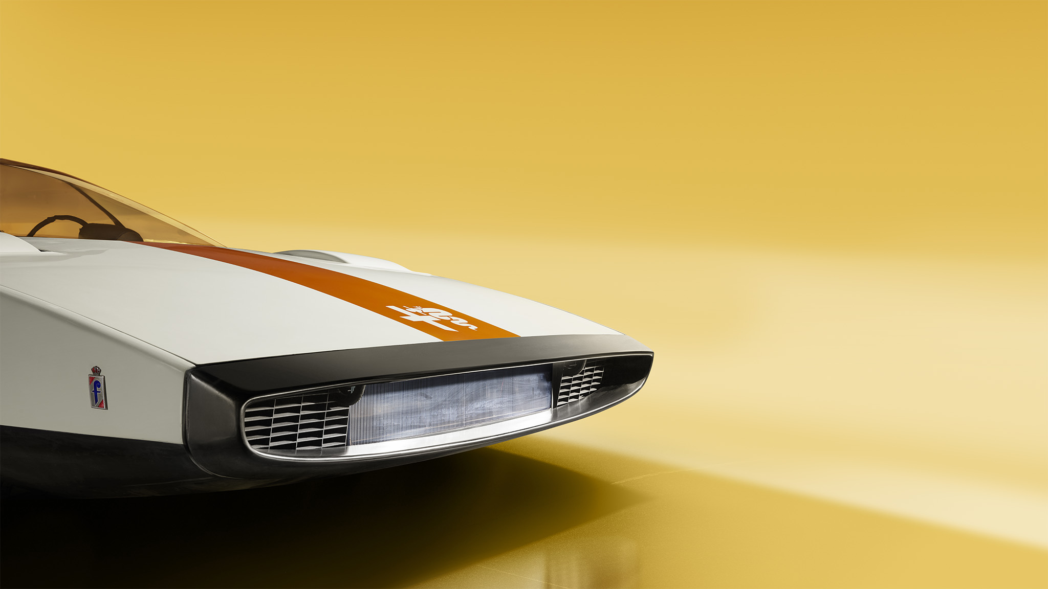 Close-up view of the front of a white sports car with an orange stripe down the center, set against a bright yellow background. The Alfa Romeo 33's futuristic design features a wide, rectangular grille with horizontal slats and sleek, aerodynamic contours—as if straight from Magneto magazine.