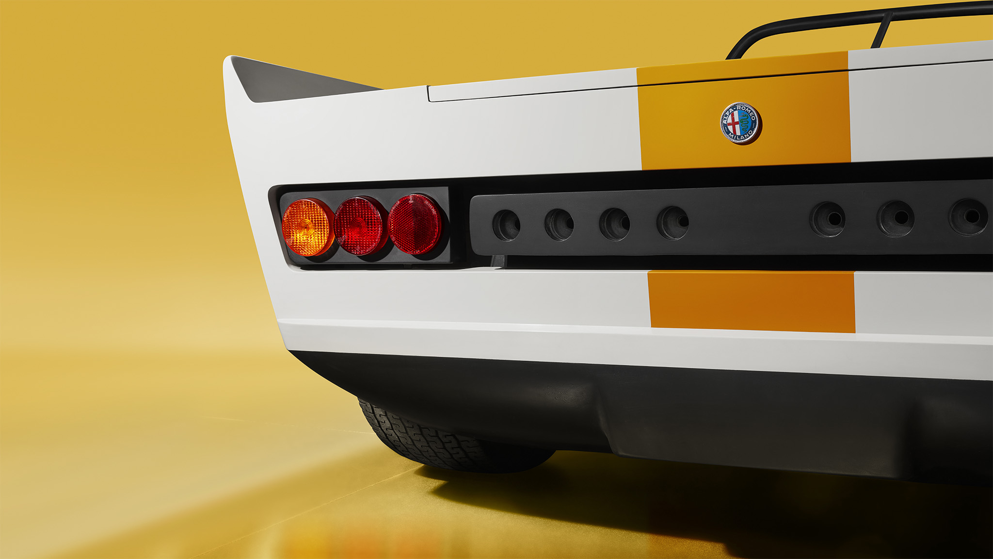 Close-up of the rear of an Alfa Romeo 33 vintage sports car with a white and orange striped design. The focus is on the left taillight cluster, which includes circular red and amber lights. The solid yellow background highlights the car's distinctive rear features, worthy of any Magneto magazine cover.