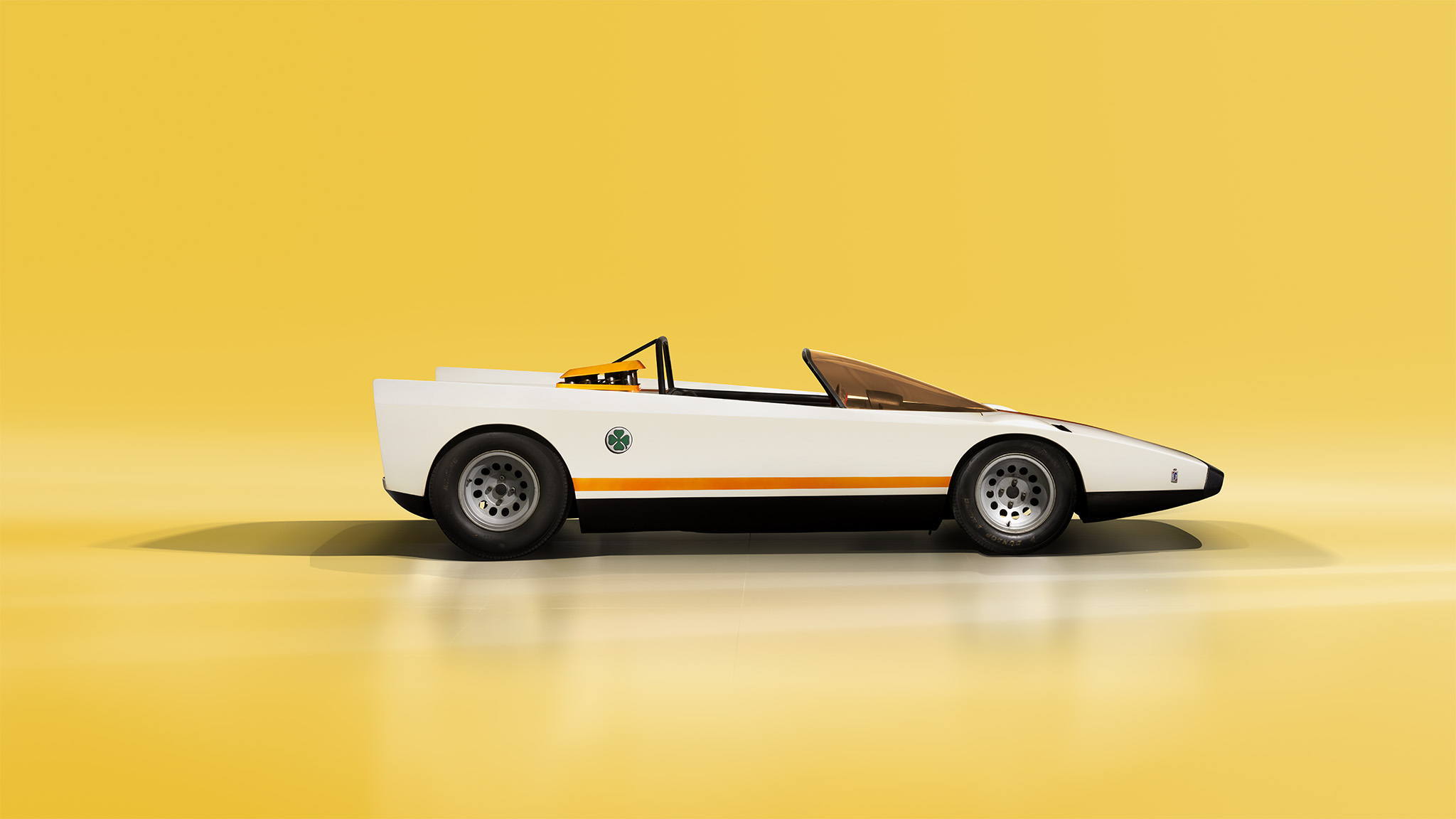 A sleek, vintage white Alfa Romeo 33 sports car with an open-top design is photographed against a vibrant yellow background. The car features angular lines, large circular wheel arches, and minimalistic design, with a single orange stripe running along its side. Perfect for the pages of Magneto Magazine.